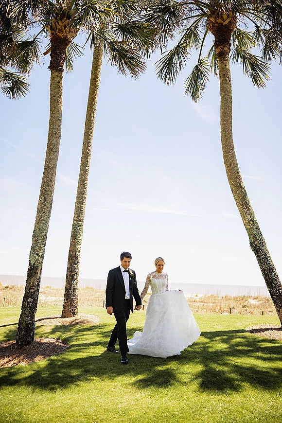 Couple portrait of bride and groom walking hand in hand, bride lifting lace long-sleeve gown train on a palm-lined lawn by the ocean