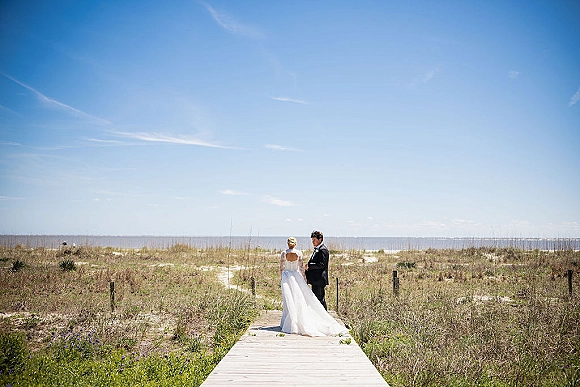 Couple portrait of beach wedding couple from behind, bride in veil and wedding dress train beside groom in tuxedo on a boardwalk by ocean horizon