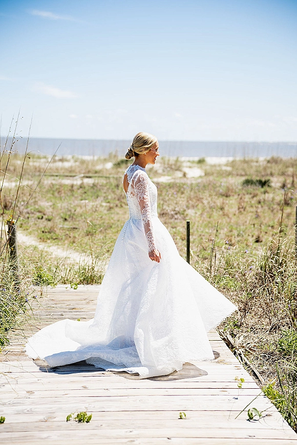 Bridal portrait of a bride in a long-sleeve lace wedding dress holding her train on a boardwalk with ocean and dunes behind her