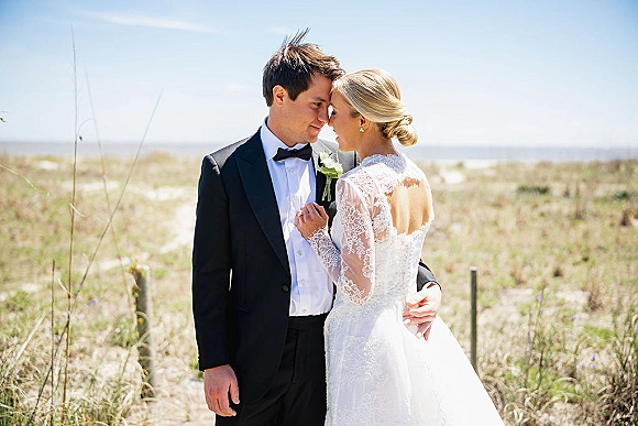 Couple portrait of bride and groom embrace, forehead touching as she holds his lapel, with ocean horizon behind beach dunes