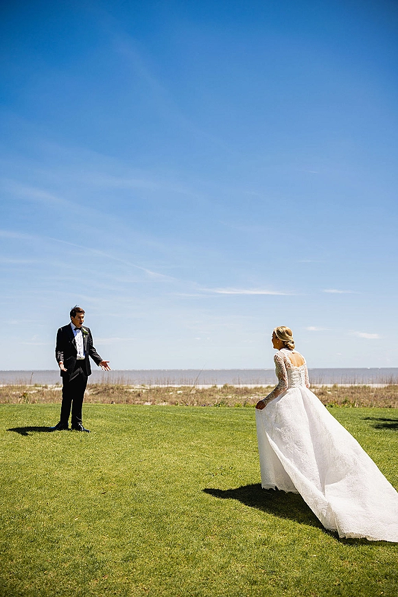 First look moment as the bride in a lace long-sleeve open-back gown and veil approaches the groom in a tux on an oceanfront lawn