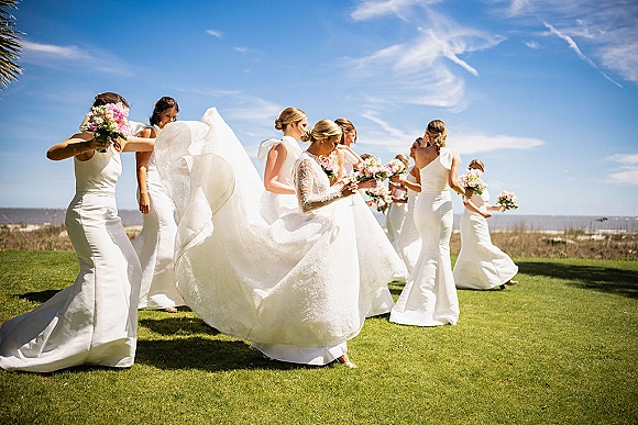 Bride with bridesmaids in a bridal party portrait, holding pink and white bouquets as her veil blows on a sunny oceanfront lawn