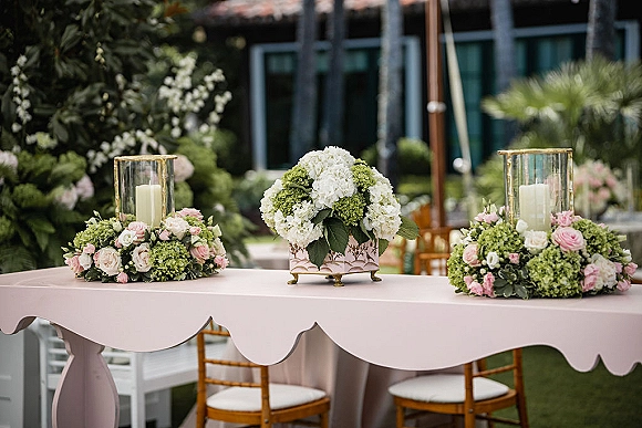 Reception sweetheart table with pink tablecloth and hydrangea centerpiece, candles and rose greenery accents on a garden lawn with palm trees