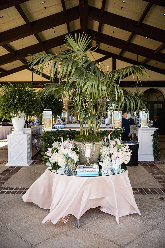 Reception cocktail decor featuring a blush-draped mirrored table with potted palm, floral vases, swan figurine and lanterns in a covered pavilion bar area