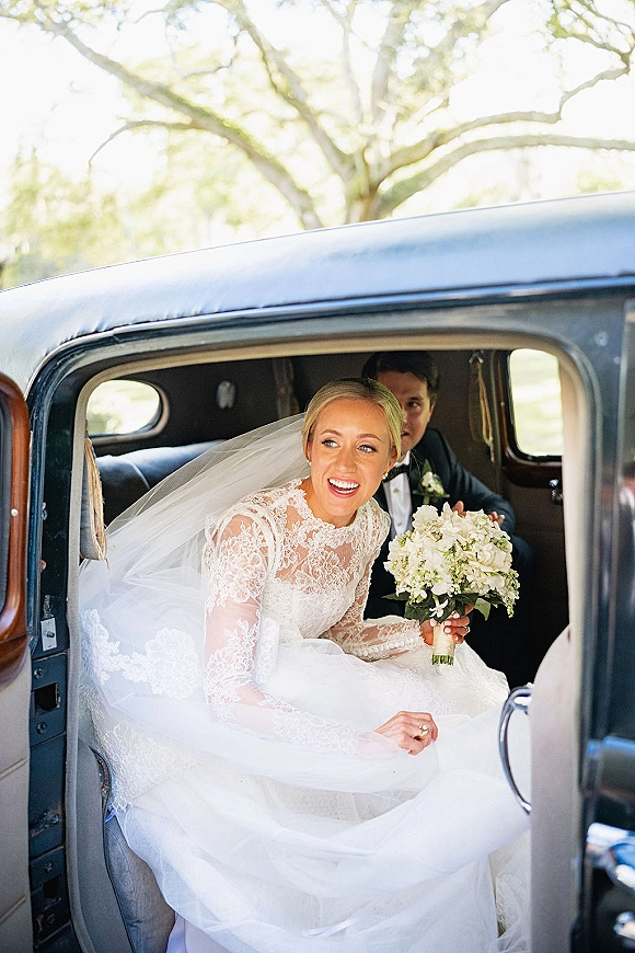 Bride portrait of a smiling bride in vintage car, holding a white hydrangea bouquet in lace sleeves by an open door in daylight
