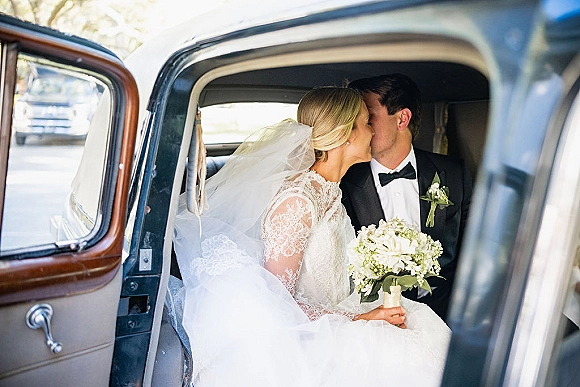 Wedding kiss between newlyweds in car, bride in lace dress and veil holding bouquet as groom in tuxedo leans in by open door