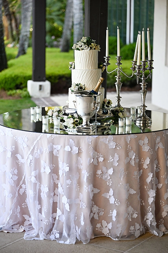 Wedding cake table with a tiered white wedding cake display, silver candelabras and champagne set on a mirrored tabletop outdoors