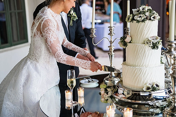 Wedding cake cutting as bride in lace sleeves and groom in tuxedo slice a tiered cake with floral topper beside silver candelabra