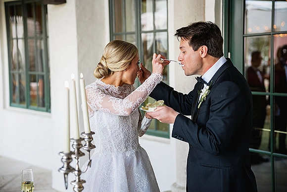 Wedding cake feeding as the couple feeding cake smiles over a slice with champagne flute and candlelit candelabra by a windowed wall