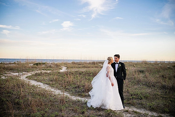 Couple portrait of bride and groom on beach dunes, her veil blowing as they gaze at each other, ocean and clouds behind them