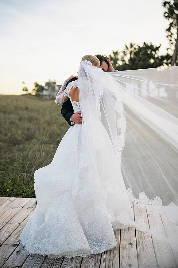 Wedding couple portrait of bride and groom embrace on a wooden deck, her long veil blowing behind a lace long-sleeve gown in a field setting