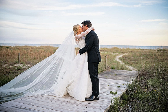 Wedding kiss portrait of bride and groom kissing on a wooden boardwalk, her long veil blowing in wind with ocean dunes behind