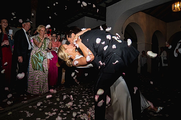 Wedding kiss moment as the couple shares a dip kiss wedding photo amid rose petal confetti, guests cheering in an arched indoor hallway.