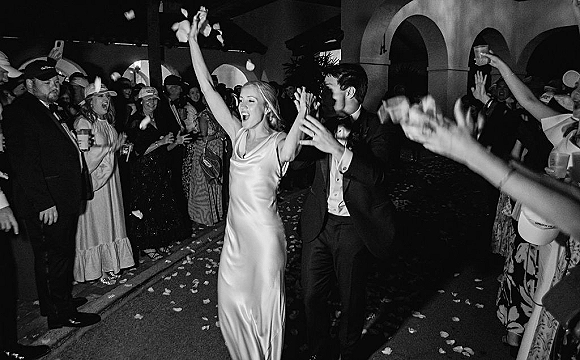 Wedding exit as bride in a satin gown and groom in tux walk through cheering guests tossing rose petals and confetti outside arched entrance at night