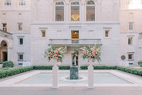 Ceremony entrance decor with floral urn wedding decor on pedestals, pink and peach blooms with greenery by a fountain in a stone courtyard