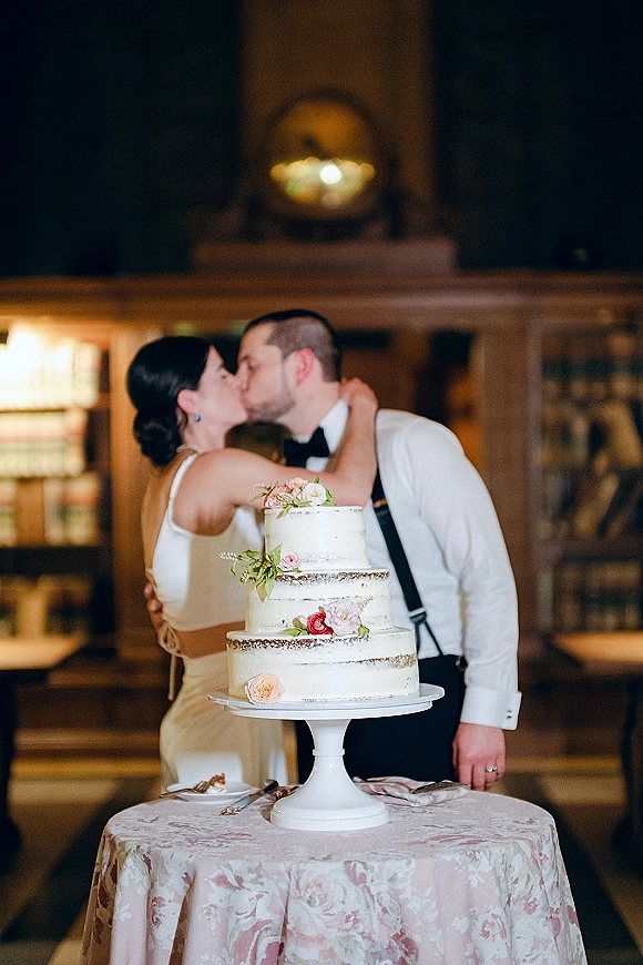 Wedding cake cutting as bride and groom share a kiss beside a three-tier buttercream cake with floral topper under warm indoor reception lighting