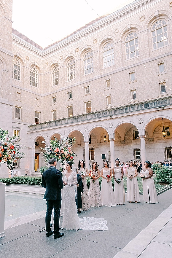 Ceremony moment at an outdoor courtyard wedding ceremony with bride in lace dress and groom in tux under floral arch, with lanterns and string lights