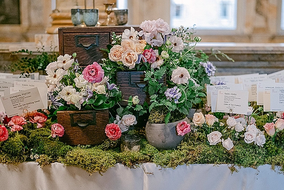 Escort card display with wedding escort cards tucked into wooden drawers, moss runner and pastel florals, set on a table by window light indoors