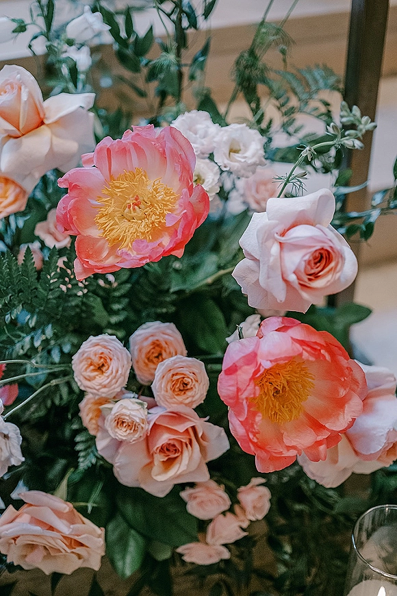 Wedding centerpiece flowers with peonies and roses in blush, coral, and white, lush greenery and fern foliage, beside a glass votive candle