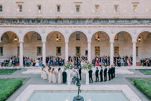 Wedding ceremony in a stone courtyard with floral ceremony altar under an arch, guests seated by a reflecting pool and fountain statue
