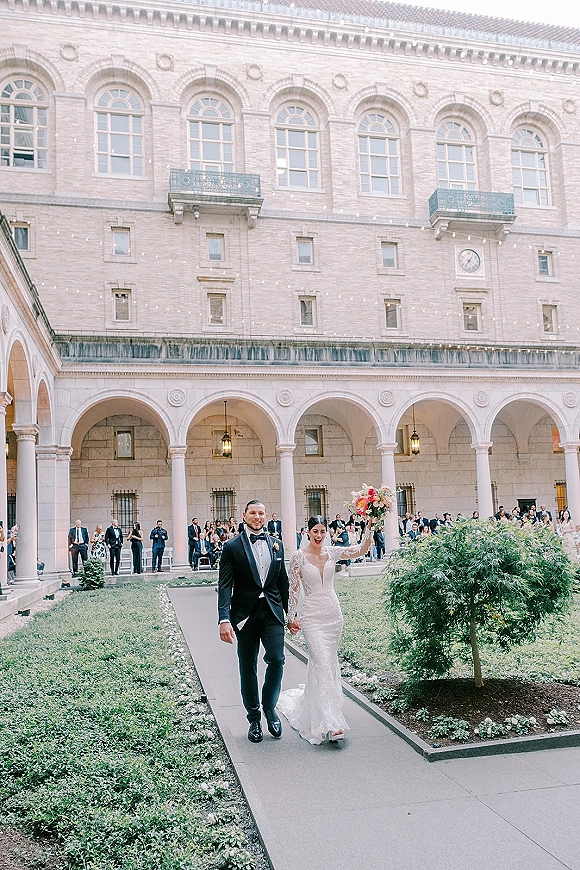 Just married exit as bride and groom walk hand in hand, bride raising bouquet under string lights in arched courtyard with guests cheering