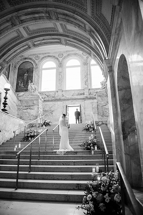 Wedding first look as the groom waiting at doorway watches the bride in a long veil and train descend a grand marble staircase with candles