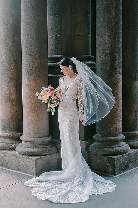 Bridal portrait of bride holding bouquet, wearing lace long sleeve wedding dress and cathedral veil by stone columns and floor