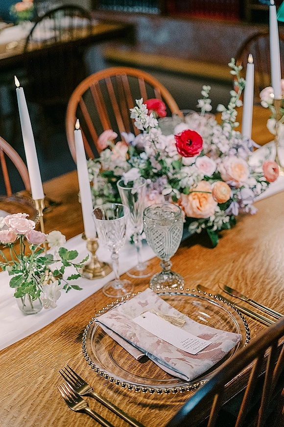 Reception tablescape with a wedding table centerpiece of pink and red florals, taper candles in brass holders, and glass chargers on a wood table in a dim venue