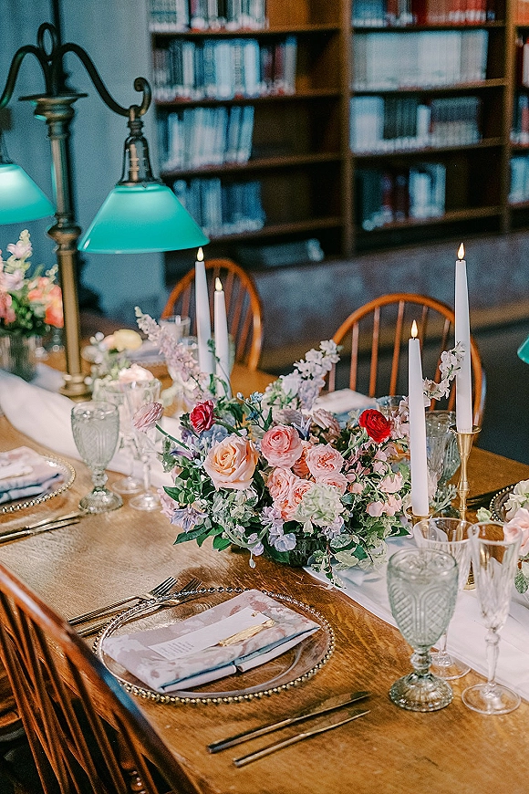 Reception tablescape with wedding table centerpiece of pastel florals, taper candles in brass candlesticks, set on a long wooden table in a library with bookshelves