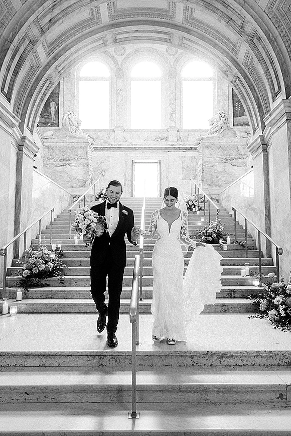 Wedding recessional as bride and groom walk down a grand marble staircase, bride holding a bouquet with veil flowing behind