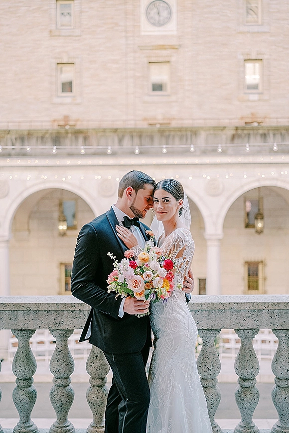 Couple portrait of bride and groom embrace on a stone balcony, her pink and peach bouquet and veil framed by arched courtyard lights