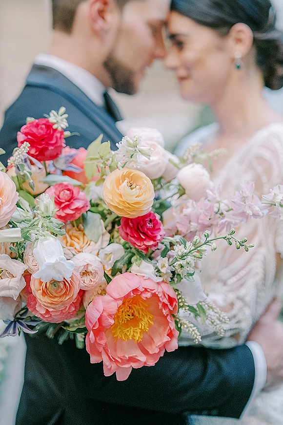 Wedding bouquet of coral and pink peonies, ranunculus, and roses with greenery held by a blurred couple against an indoor wall
