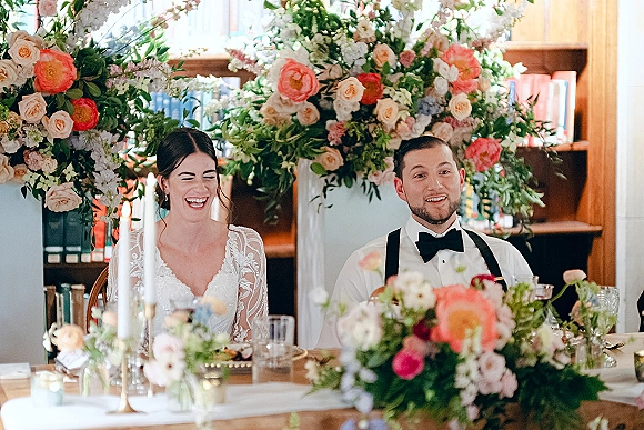 Wedding sweetheart table with bride and groom at table, framed by tall florals and candles against library bookshelves, wood paneling
