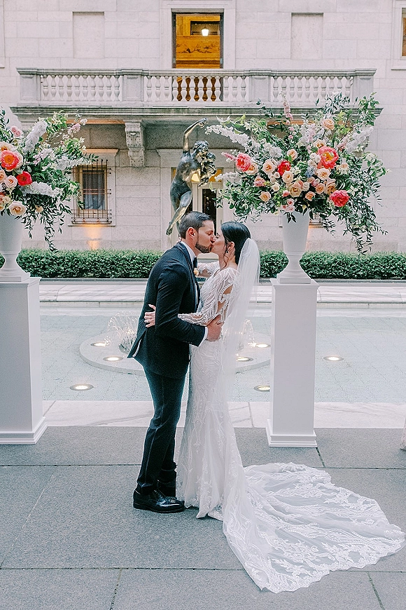 Wedding kiss portrait of bride and groom kissing beneath a flowing veil, framed by grand floral urns and a courtyard fountain backdrop
