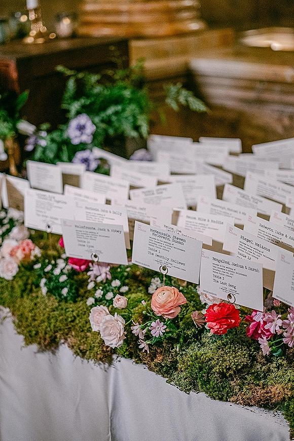 Wedding escort cards on clips in an escort card display, arranged over a moss runner with floral accents against a wood wall and greenery backdrop