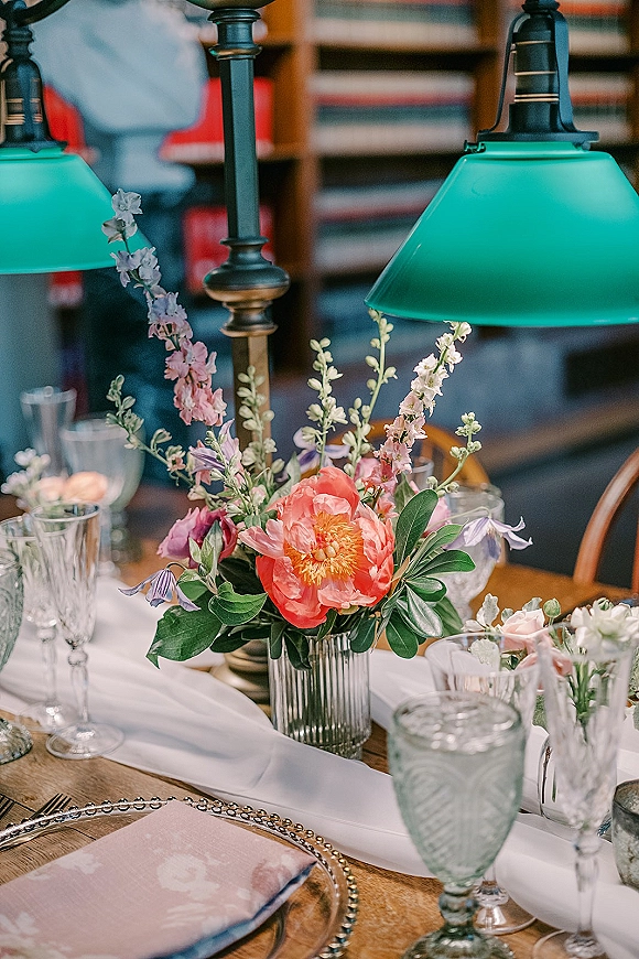 Reception tablescape with a wedding table centerpiece of peonies and pastel florals in a ribbed vase, candles and green lamps on a wood table by bookshelves