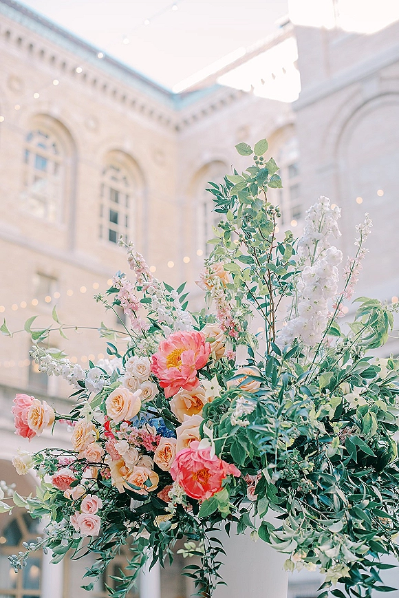 Wedding floral arrangement with lush greenery, roses, peonies, and pastel blooms beneath string lights in an airy courtyard with arched windows