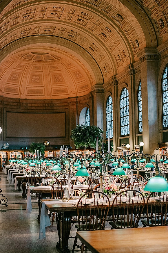 Reception tablescape in a library wedding reception with long white-linen banquet tables, green banker lamps, taper candles, and bud vase florals under arched ceiling