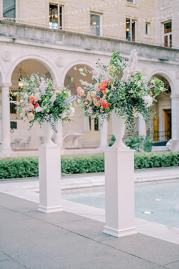 Wedding floral pillars with ceremony entrance flowers in pedestal urns on white columns, framed by an arched courtyard and reflecting pool