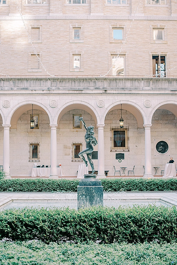 Wedding venue courtyard with string lights over cocktail tables, featuring a central fountain and statue beneath stone arches and columns