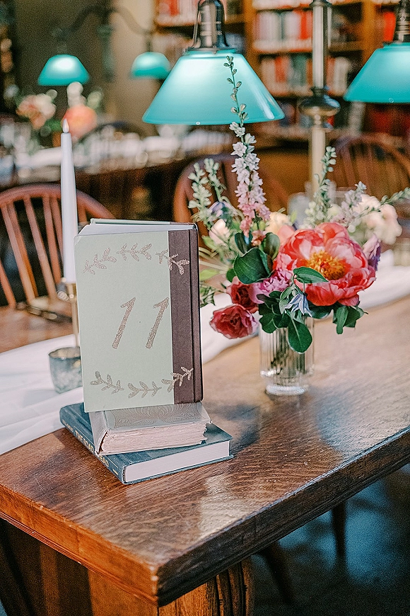 Wedding table number display with book table numbers, calligraphy card on stacked books beside florals in a glass vase and teal lamp on wood table