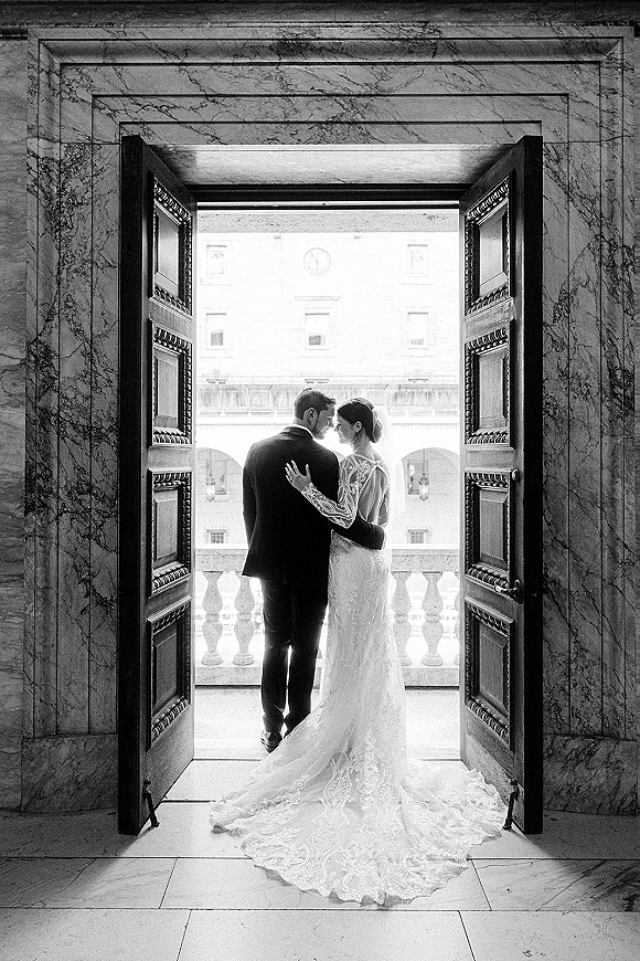Couple portrait of bride and groom from behind in a doorway, her cathedral veil and long lace train flowing toward a marble balcony view