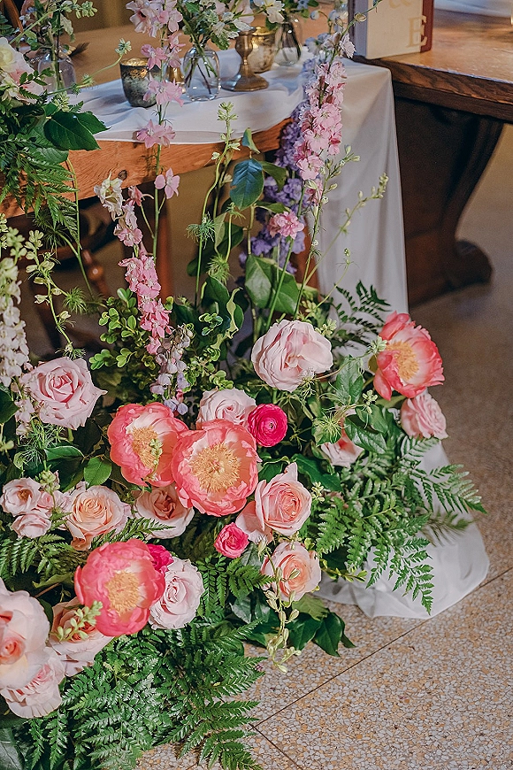 Wedding floral arrangement with pink roses and coral peonies in a brass vase on a wooden table with bud vases and votive candles near a bar counter