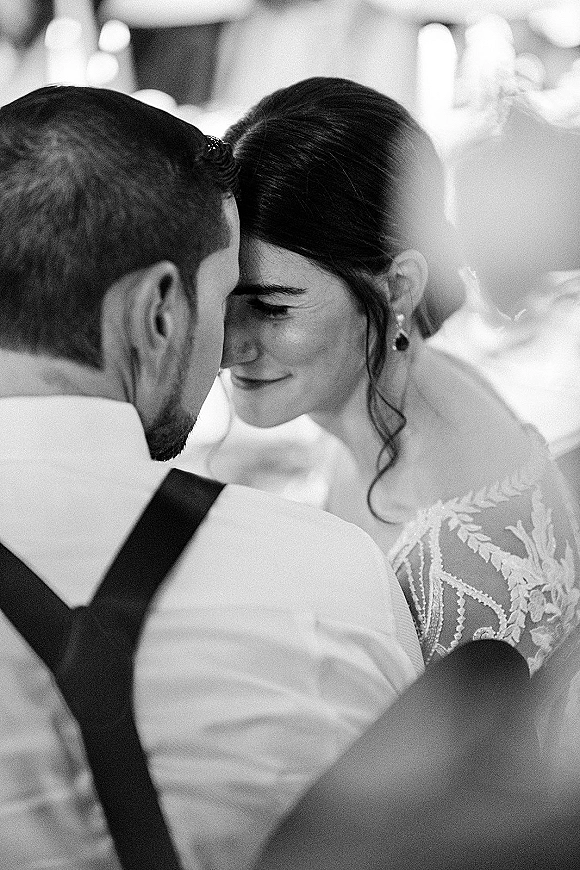 Couple portrait in a black and white wedding photo, bride and groom touching foreheads as he embraces her amid blurred reception tables and guests