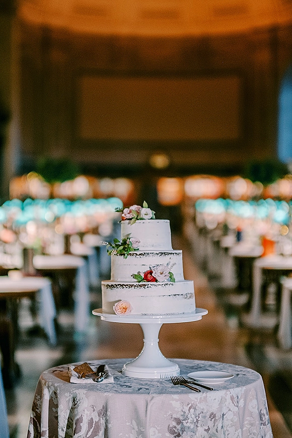 Wedding cake with white buttercream frosting on a stand, topped with fresh flowers, with cake knife and server on lace tablecloth in reception hall