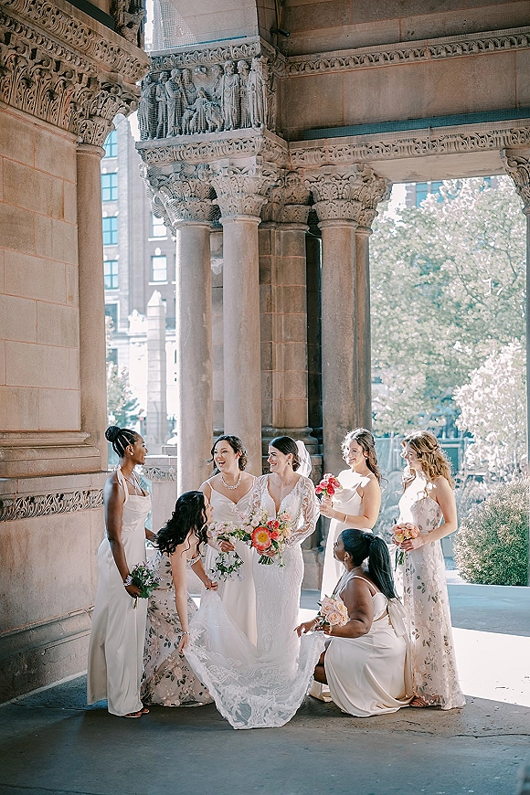 Bride with bridesmaids smiling in mismatched dresses, holding bouquets beside a carved stone archway and columns outdoors