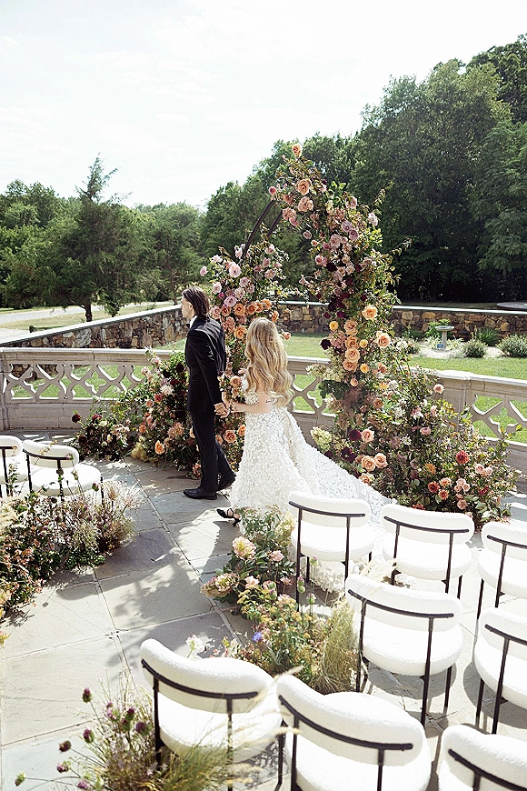 Ceremony setup with outdoor ceremony arch featuring peach and pink flowers and greenery, white chairs lining a stone terrace aisle