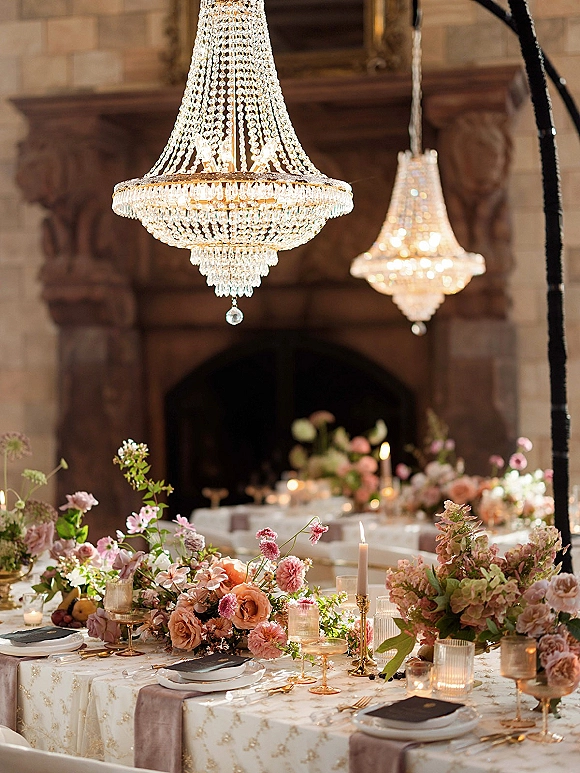 Reception tablescape with wedding table centerpieces, blush florals and taper candles under crystal chandeliers beside a stone fireplace