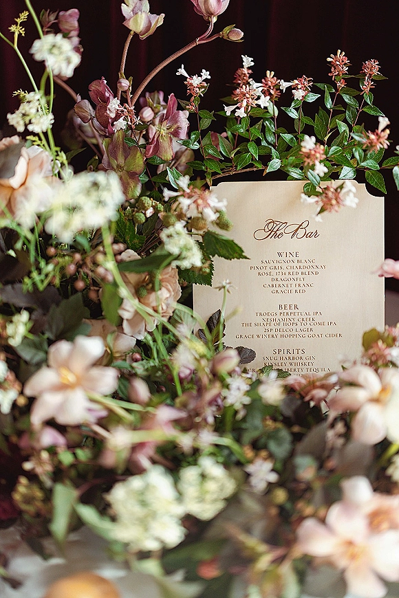 Wedding bar menu and signature drinks menu card in classic script, framed by pink and white flowers with greenery against a dark backdrop
