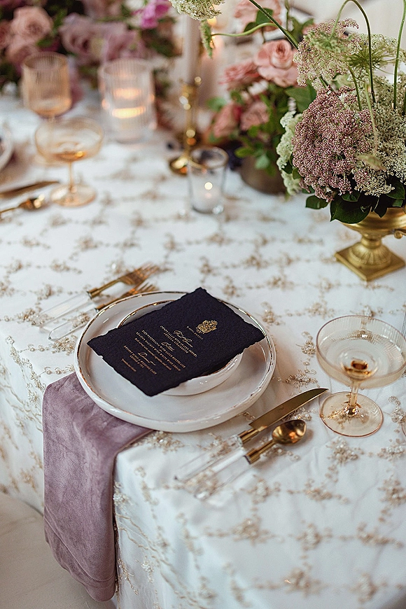 Reception tablescape with wedding place setting, floral centerpiece in compote vase, taper and votive candles, champagne coupes on embroidered tablecloth